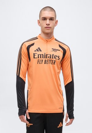 Ung mand iført orange og sort langærmet Adidas sportstop med "Emirates Fly Better" og Arsenal fodboldklub logoer.