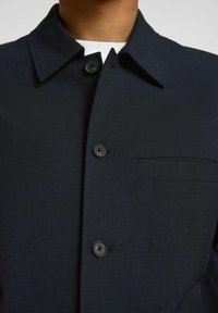 Giacca blu navy con bottoni, dotata di colletto, realizzata in un tessuto testurizzato, con cinque bottoni e una tasca sul petto.