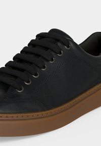 Baskets en cuir noir avec des lacets noirs et des œillets métalliques, dotées d'une semelle en caoutchouc marron sur un fond clair et uni.