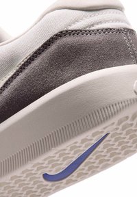 Gros plan du talon d'une basket beige avec un détail en daim gris et un swoosh Nike violet sur une semelle en caoutchouc texturée.