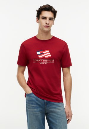 Νεαρός άντρας που φοράει κόκκινη μπλούζα Tommy Hilfiger με σχέδιο σημαίας και μπλε τζιν, στέκεται μπροστά σε ένα απλό λευκό φόντο.