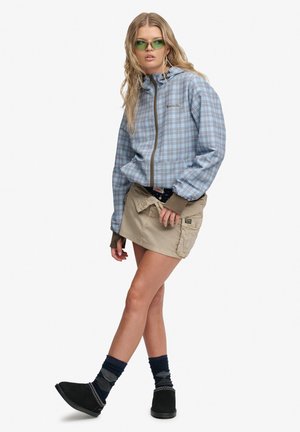 Jeune femme portant des lunettes de soleil vertes, une veste zippée à carreaux bleus, une mini-jupe beige, des chaussettes foncées et des chaussures noires, debout avec une jambe croisée.