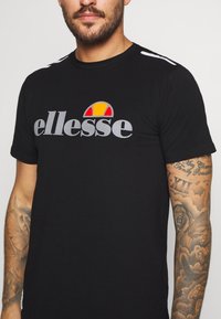 Black cotton t-shirt featuring a large grey "ellesse" logo with a colorful sun graphic above. White accents on shoulders. Classic fit.