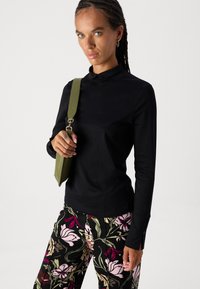Schwarzes Langarmoberteil mit einem Stehkragen und seitlichen Schlitzen, kombiniert mit Blumenmuster-Hosen in Rosa- und Grüntönen sowie einem olivgrünen Tragriemen.