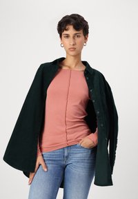 Chemise en velours côtelé vert foncé, ouverte sur un haut à manches longues rose clair avec couture centrale, associée à un jean bleu.