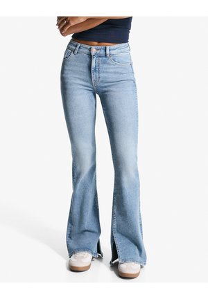 Femme portant un jean flare taille haute bleu clair avec des fentes à l'avant, associé à des baskets blanches et un top court foncé, bras croisés.