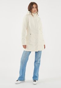 Cappotto lungo crema con cappuccio, chiusura con zip, tasche laterali e dettagli elasticizzati. Indossato sopra jeans azzurri chiari e sneaker.