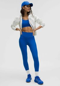 Mėlyna sportinė liemenėlė, aukšto juosmens mėlynos leggins, baltas užtrauktukas striukė, mėlyna kepurė ir mėlyni sportiniai batai; glotni tekstūra, prigludęs dizainas.