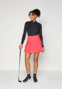 Fekete hosszú ujjú felső és ráncolt élénk rózsaszín golf szoknya. A modell golfütőt tart, fekete tornacipőt és zoknit visel. Egyszínű háttér.