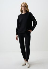 Zwarte sweatshirt met lange mouwen en een relaxte pasvorm, gecombineerd met bijpassende zwarte joggers met witte zijstreepjes. Witte sneakers zichtbaar.