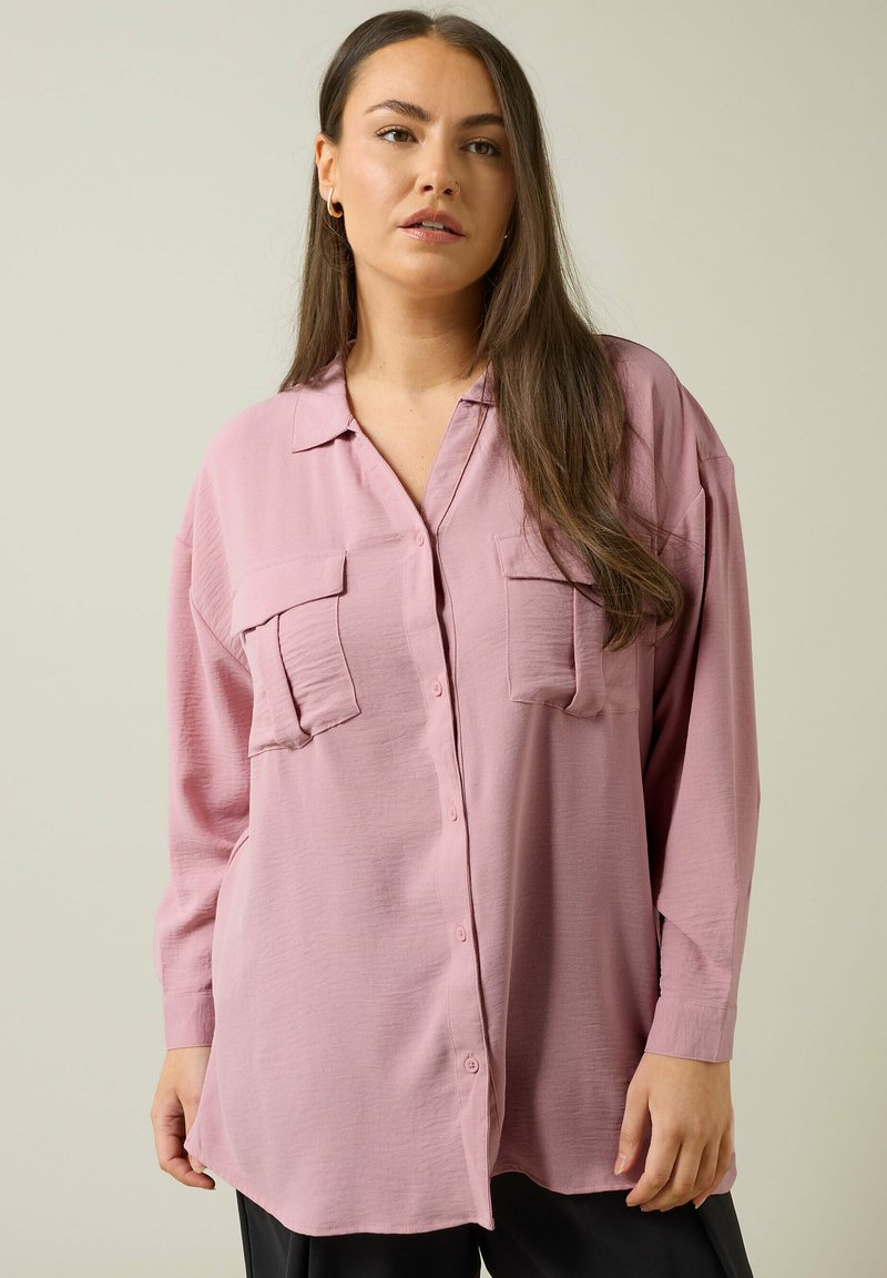 Camisa de manga larga rosa suave con botón en la parte delantera, dos bolsillos en el pecho y un corte relajado. Hecha de una tela ligera y texturizada.