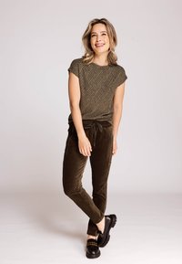 Kurzärmeliges Oberteil mit olivgrünem Muster, kombiniert mit dunkelgrünen Cordhosen und schwarzen Schuhen mit goldenen Akzenten, vor einem neutralen Hintergrund stehend.