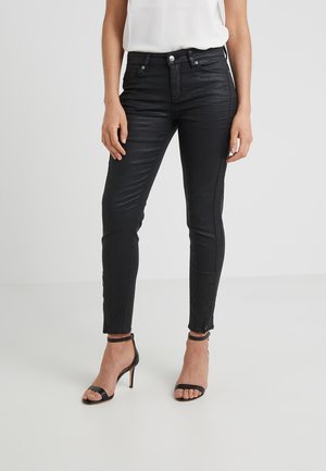 Vrouw draagt nauwsluitende zwarte broek en zwarte hoge hakken sandalen, staand tegen een effen lichte achtergrond.