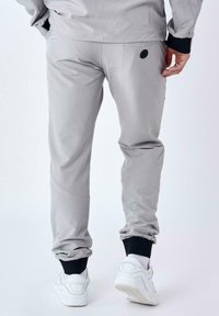 Pantalones jogger de color gris claro con puños elásticos, bolsillos laterales y un acento de logo negro. Textura suave con un diseño estilizado.