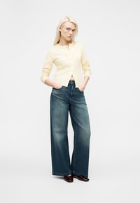 Femme aux longs cheveux blonds portant un cardigan boutonné jaune clair et un jean large bleu, debout les mains dans les poches sur un fond blanc.