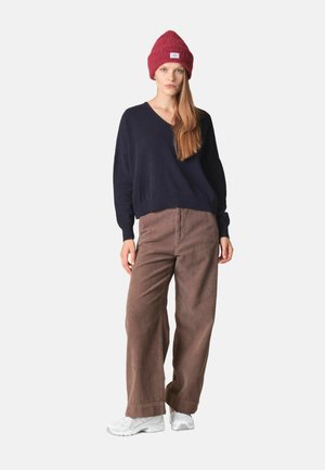 Femme portant un bonnet rouge en maille, un pull noir ample à col en V, un pantalon marron large en velours côtelé et des baskets blanches, debout sur un fond uni.