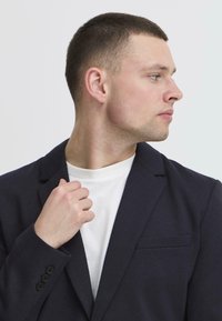Blazer bleu marine foncé avec un revers structuré, une poche poitrine simple et un détail à trois boutons aux poignets, porté sur un t-shirt blanc à col rond.