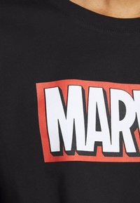 Sort skjorte med stort hvidt "MARVEL" tekst omkranset af rødt, båret af en person med synlig nakke og skulderområde.