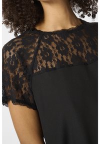 Femme portant une robe noire avec des manches courtes en dentelle florale et un empiècement en dentelle autour du décolleté, laissant apparaître la peau sous les détails de dentelle.
