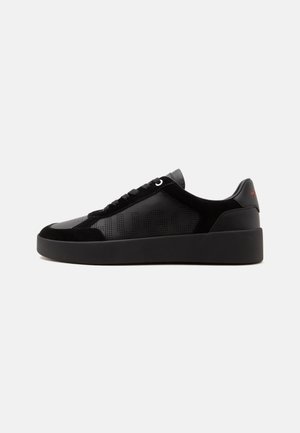 Baskets basses noires avec semelle épaisse, panneau latéral perforé affichant le nom de la marque, design à lacets et logo rouge subtil sur l'onglet du talon.