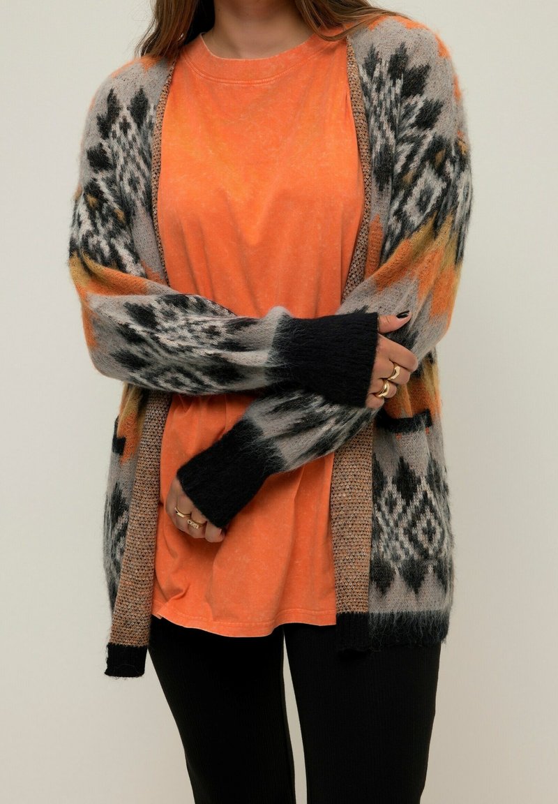 Personne portant une chemise orange et un long cardigan duveteux à motifs géométriques noirs, gris et orange, bras croisés à la taille.