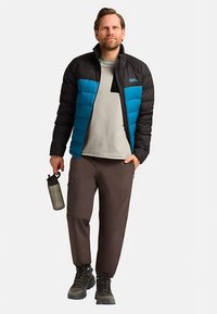 Donsjack voor heren met een zwarte bovenkant en blauwe onderkant, gecombineerd met een grijs T-shirt, bruine broek en grijze wandelschoenen. Houdt een fles water vast.