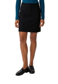 Zwarte kokerrok met een getailleerde pasvorm, voorzien van zijzakken en een gladde stofafwerking. Gecombineerd met glanzende zwarte loafers.
