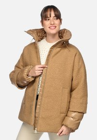 Chaqueta de gran tamaño en tono marrón con un exterior de lana texturizada, mangas de plumas brillantes, un cuello alto y cierre de cremallera con bolsillos laterales.