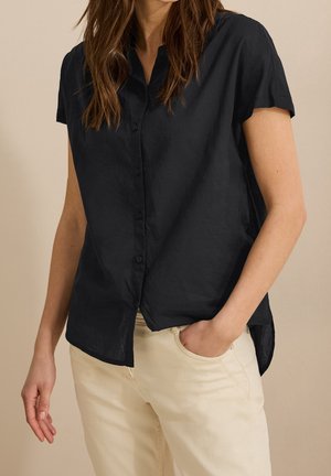 Woman wearing a short-sleeve black button-up shirt with a collar and light beige pants, one hand in pocket, against a neutral background.
