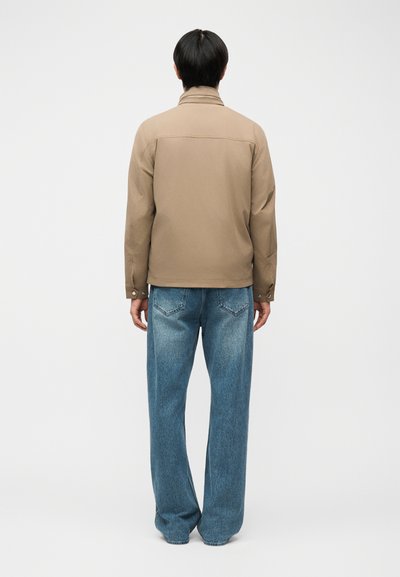 Homme debout, de dos, portant une veste beige et un jean bleu ample, sur un fond clair uni.