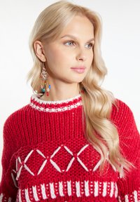 Czerwony sweter z dziewiarskim wzorem w białym kolorze, z wysokim dekoltem. Założony z długimi, kolorowymi kolczykami z koralików, prezentującymi różne kształty.