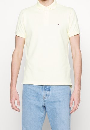 Mand iført en lys gul kortærmet polo og lyseblå jeans mod en ensfarvet hvid baggrund.