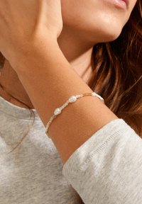 Gold-akzentuertes Armband mit weißen Perlen und kleinen Perlen in verschiedenen Farbtönen, getragen an einem Handgelenk mit einem weichen grauen Pullover.