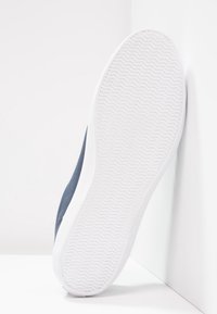 Semelle en caoutchouc blanche d'une basket bleue avec motif cranté en zigzag, présentée debout contre un mur d'angle blanc.