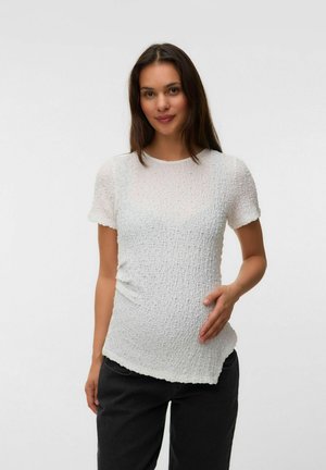 Femme enceinte aux longs cheveux bruns, portant un haut blanc texturé à manches courtes et un pantalon noir, tenant son ventre et souriant à la caméra.