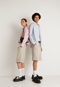 Kaks noort täiskasvanut seisavad seljaga üksteise poole, kandes triibulisi nööbiga särke, beeže põlvpükse, valgeid sokke ja musti kingi valge taustaga.