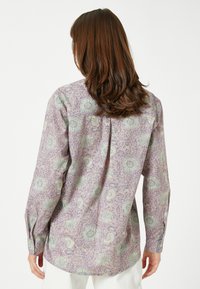Lange mouw shirt van lichtpaarse stof met bloemmotieven in groen en geel. Heeft een rechte rugloop en knoopmanchetten.
