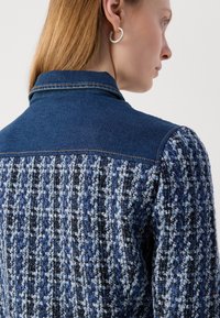 Veste en denim avec un col bleu foncé, tissu tissé texturé en bleu et noir, et une couture visible le long de l'épaule.