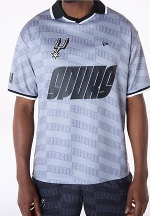 Mężczyzna ubrany w szarą koszulkę San Antonio Spurs oraz dopasowane spodenki z powtarzającym się napisem „San Antonio” i logo drużyny na piersi.