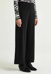 Pantaloni neri a gamba larga con una texture liscia, caratterizzati da risvolto e abbinati a stivaletti neri borchiati.