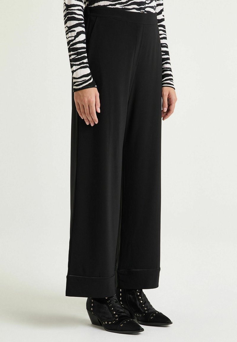 Pantaloni neri a gamba larga con una texture liscia, caratterizzati da risvolto e abbinati a stivaletti neri borchiati.