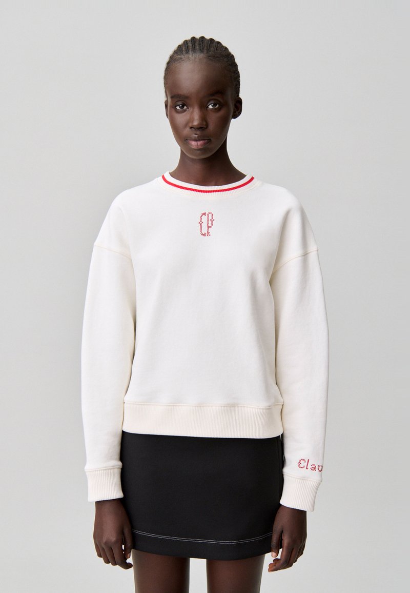 Sweat-shirt crème avec encolure côtelée rouge et logo brodé sur la poitrine. Porté avec une jupe noire présentant des détails de couture contrastants.