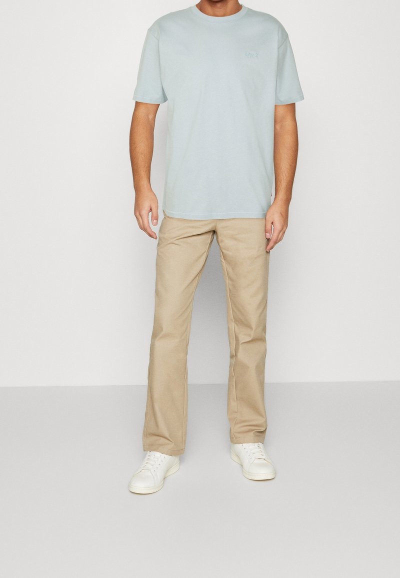 Homme portant un t-shirt bleu clair à manches courtes, un pantalon beige droit et des baskets blanches, debout devant un fond blanc uni.