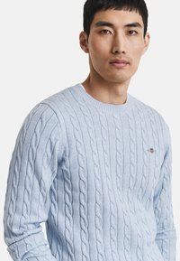 Himmelblauer Kabelstrick-Pullover mit klassischem Rundhalsausschnitt und gerippten Bündchen. Kleines gesticktes Detail auf der Brust.