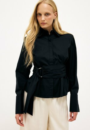 Blonde Frau trägt ein schwarzes Langarmshirt mit breiten Manschetten und einem breiten Taillengürtel, kombiniert mit hellbeigen Hosen, vor hellem Hintergrund.