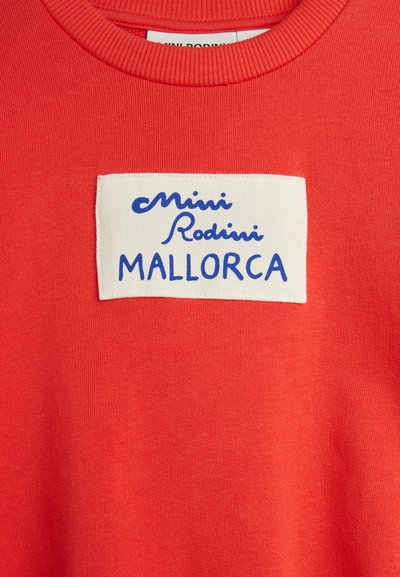 Piros kötött anyag fehér téglalap alakú cimkével, amelyen kék dőlt és nyomtatott betűkkel az állati rész közelében a "Mini Rodini MALLORCA" felirat olvasható.