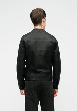 Homme portant une veste en cuir noire et un jean noir, debout de dos sur un fond blanc uni.