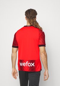 Maglia sportiva rossa con strisce orizzontali nere, con rifiniture nere sul colletto e sulle maniche. Testo "wefox" sulla parte bassa della schiena.