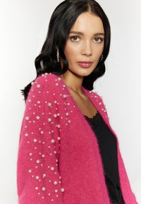 Cárdigan rosa con adornos de perlas en las mangas, hecho de un material suave y texturizado. Superpuesto sobre una blusa de encaje negra.