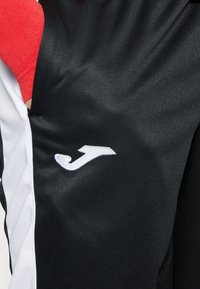 Svart idrottströja med vita accenter och en röd ärm, med en framträdande vit logotyp på framsidan, tillverkad av ett smooth, lätt material.